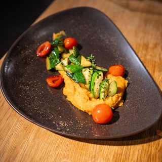 Ein modernes Gericht bestehend aus einer Kugel Süßkartoffelpüree, garniert mit sautierten Zucchini, Kirschtomaten und einem Zweig frischem Thymian, präsentiert auf einem stilvollen schwarzen Teller auf einem Holztisch.