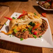 Ein mit Gemüse gefülltes Blätterteiggebäck wird auf einem weißen Teller serviert, dazu gibt es pürierte Bohnen und garniert mit frischen Kräutern und geschnittenen roten Paprikaschoten. Das Gericht wird auf einem Holztisch präsentiert.