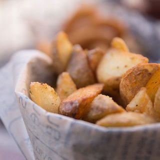 Nahaufnahme von knusprigen, goldbraunen Kartoffelspalten, die in Zeitungspapier eingewickelt sind. Die Kartoffeln sind mit Kräutern und Gewürzen gewürzt, wodurch ihre strukturierte Schale und ihr weiches Inneres hervorgehoben werden. Der Hintergrund ist leicht verschwommen und lenkt die Aufmerksamkeit auf den appetitanregenden Snack.