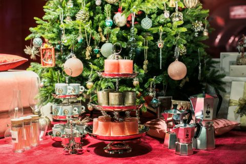 Eine festliche Dekoration vor einem Weihnachtsbaum mit bunten Kerzen auf mehreren Etagen, Tassenstapeln, Kaffeemaschinen und dekorativen Ornamenten. Die Atmosphäre ist lebendig mit warmen Rot- und Grüntönen.