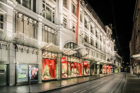Ein hell erleuchtetes Kaufhaus bei Nacht, geschmückt mit Weihnachtsdekorationen. Das Gebäude verfügt über große Fenster, die festliche Szenen mit roten Akzenten und Weihnachtsbäumen zeigen. Die Straße ist leer und die Fassade leuchtet mit dekorativen Lichtern.