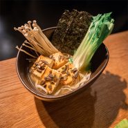 Eine Schüssel Ramen mit Tofu, Enoki-Pilzen, gehackten Frühlingszwiebeln, einem Blatt Seetang und Pak Choi obendrauf, auf einem Holztisch platziert.