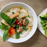 Eine Schüssel Nudelsuppe mit geschnittenen Zwiebeln, Karotten, Fleischstücken und roten Chilischoten, garniert mit frischen Kräutern. Ein Beilagenteller mit Gemüse steht neben der Schüssel auf einem Holztisch.