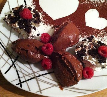 Ein Teller mit drei Kugeln Schokoladenmousse, garniert mit frischen Himbeeren. Darauf liegen zwei Kleckse Sahne, garniert mit Schokoraspeln. Der Teller ist mit herzförmigen Motiven aus Kakaopulver verziert.