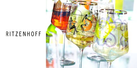 Un ensemble de verres Ritzenhoff colorés et artistiques, remplis de boissons. Chaque verre a un design unique avec des motifs et des figures vibrantes. L'arrière-plan présente des reflets de lumière et d'autres verres, créant une atmosphère festive.