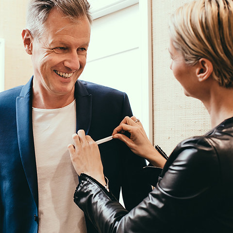 Un homme souriant en costume bleu se fait remettre en place par une femme aux cheveux courts et portant une veste en cuir. Elle ajuste le revers de son veston, tandis qu'il a l'air satisfait. Ils se trouvent dans une pièce bien éclairée aux murs de couleur neutre.