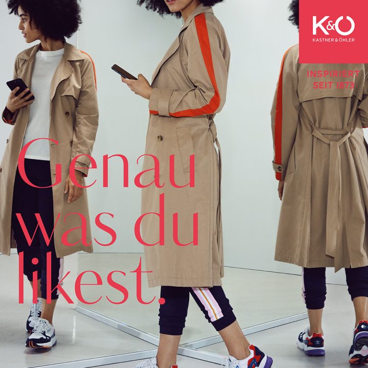 Une personne aux cheveux bouclés, portant un trench-coat beige et des baskets, regarde son téléphone portable dans un espace moderne avec des miroirs. Le texte est « Exactement ce que tu aimes ». En haut à droite, on voit un carré rouge avec « Kastner & Öhler ».