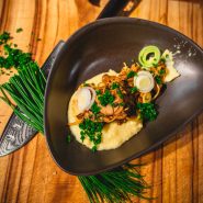 Ein schwarzer Teller mit cremigem Kartoffelpüree, garniert mit sautierten Pilzen, geschnittenem Lauch und gehackten Kräutern. Das Gericht steht auf einer Holzoberfläche, ist mit grünem Schnittlauch garniert und wird von einem gemusterten Messer begleitet.