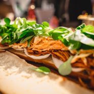 Nahaufnahme eines belegten Sandwichs mit geraspelten Karotten, Gemüse und Zwiebelscheiben auf einer Brotscheibe. Der Fokus liegt auf den frischen Zutaten, der Hintergrund ist unscharf.