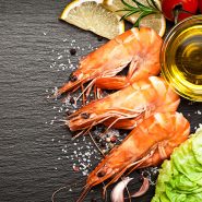 Drei gekochte Garnelen auf einer dunklen Schieferplatte, umgeben von Zitronenscheiben, Rosmarin, Pfefferkörnern und Meersalz. Dazu eine kleine Schale Olivenöl und etwas Salat.