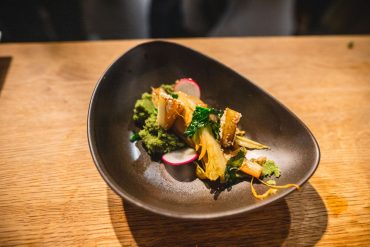 Ein Gourmetgericht wird elegant in einer dunklen, asymmetrischen Schüssel präsentiert. Es besteht aus einer Gemüsemischung, darunter Blattgemüse, Radieschenscheiben und Artischocken, kunstvoll arrangiert auf einer grünen Soße, alles auf einem Holztisch.