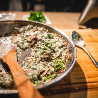 Eine Bratpfanne mit einer cremigen Mischung aus Pilzen und Erbsen steht auf einem hölzernen Schneidebrett. In der Pfanne liegt ein Holzlöffel.