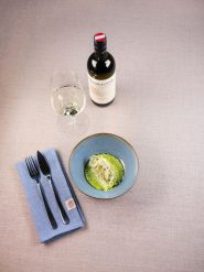 Auf einer beigen Tischdecke steht eine Suppenschüssel, garniert mit einer Scheibe Zitrusfrucht. Daneben liegen eine gefaltete blaue Serviette mit Messer und Gabel, ein Glas Weißwein und eine Flasche Wein. Das Ambiente ist elegant und schlicht.