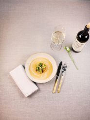Ein Gourmetgericht wird elegant auf einem beigen Teller präsentiert, begleitet von Messer und Gabel mit goldenem Griff. Daneben stehen ein Glas Weißwein und eine Weinflasche, mit einer weißen Serviette und einer einzelnen Blume auf einer hellgrauen Tischdecke.