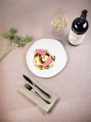 Ein Teller mit Rindfleischscheiben auf einem Bett aus Kartoffelpüree, garniert mit grüner Soße. Auf der rechten Seite stehen eine Flasche Wein und ein gefülltes Weinglas. Auf der linken Seite liegen Messer und Gabel auf einer Serviette, daneben ein Zweig Grünzeug.