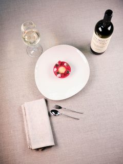 Ein weißer Teller mit einem roten Dessert, garniert mit Himbeeren und einer Kugel Eis. Daneben stehen ein mit Weißwein gefülltes Weinglas, eine Flasche Rotwein und zwei Löffel auf einer Tischdecke neben einer gefalteten Serviette.