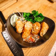 Eine schwarze Schüssel mit geschnittenen Frühlingsrollen, gefüllt mit Gemüse, garniert mit Sojasprossen und Koriander. Daneben liegt eine Limettenscheibe. Auf dem Holztisch neben der Schüssel liegt ein schwarzes Messer.