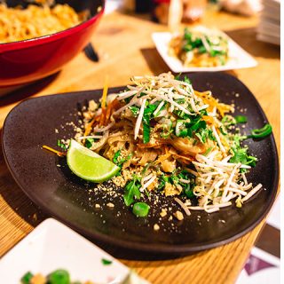 Ein Teller Pad Thai, garniert mit Sojasprossen, gehackten Erdnüssen, Frühlingszwiebeln und Koriander. Eine Limettenscheibe liegt daneben. Das Gericht steht auf einem schwarzen Teller, im Hintergrund ist eine weitere Portion zu sehen.