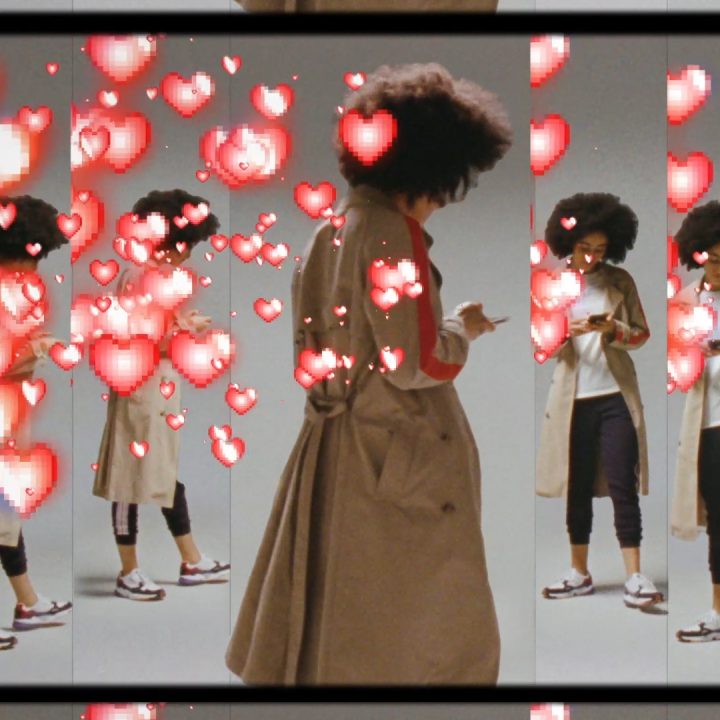 Une femme aux cheveux bouclés et portant un long manteau se tient devant un fond réfléchissant qui crée de multiples reflets. Elle tient un téléphone et des animations de cœurs rouges sont superposées à l'image, créant un effet vibrant et romantique.
