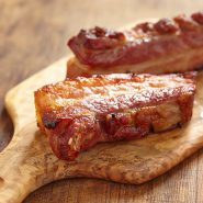 Nahaufnahme von saftigen, gebratenen Schweinerippchen mit goldbrauner Kruste auf einem rustikalen Holzschneidebrett. Die Rippchen glänzen und sind leicht verkohlt, was ihr zartes und würziges Aussehen unterstreicht.