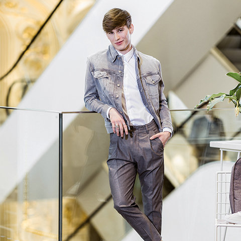 Une personne en veste en jean claire et pantalon gris est nonchalamment appuyée contre une balustrade en verre dans une pièce moderne et bien éclairée. Elle a les mains dans les poches et les cheveux courts et bruns.