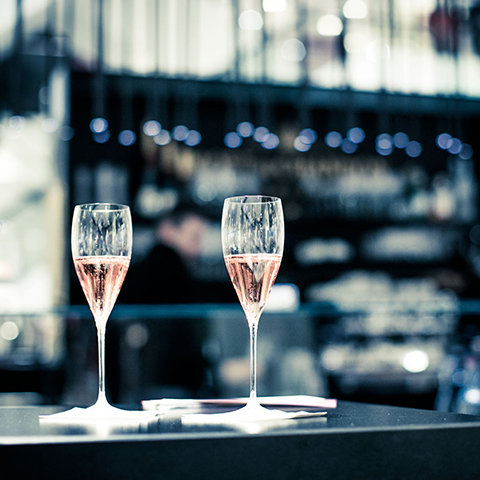 Deux coupes de champagne rosé sur une table sombre. L'arrière-plan est légèrement flou et montre un bar faiblement éclairé avec une lumière bokeh discrète. La scène dégage un sentiment d'élégance et de célébration.