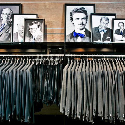 Dans la vitrine d'un magasin de vêtements, deux portants présentent des costumes gris sous des portraits encadrés en noir et blanc d'hommes portant des nœuds papillon, le tout disposé sur un fond en bois.