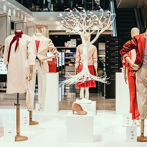 Une exposition-vente avec des mannequins portant des vêtements à la mode, y compris des tenues rouges et blanches. Entre eux se trouve une sculpture d'arbre blanche décorative, et diverses chaussures sont exposées sur des socles blancs. Un escalator est visible en arrière-plan.
