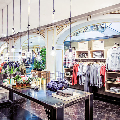 Une boutique de vêtements élégante avec des portants bien rangés remplis de chemises et de pulls. L'espace présente un décor vintage, des ampoules suspendues et des détails muraux artistiques. Une table centrale expose des vêtements pliés et des plantes en pot, créant une atmosphère chaleureuse.