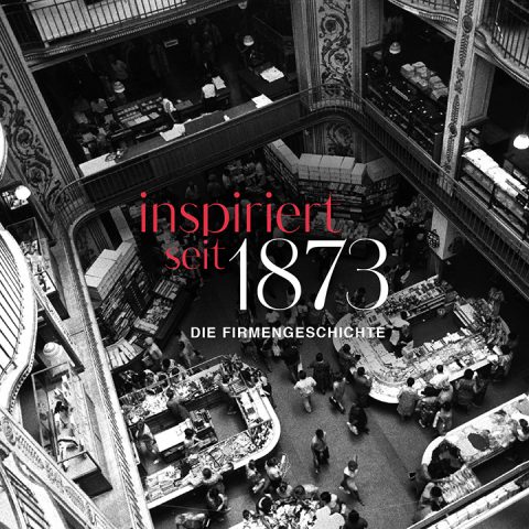 Une photo en noir et blanc d'un grand magasin animé à plusieurs étages vu d'en haut, montrant des acheteurs et divers comptoirs. Le texte en rouge et blanc indique « inspiré depuis 1873 - l'histoire de l'entreprise ».