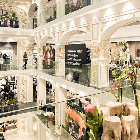 Un centre commercial à plusieurs étages avec un décor élégant en blanc et or. Les gens flânent dans les magasins et de grandes expositions d'art sont présentées aux étages supérieurs. Des balustrades en verre et des arrangements floraux contribuent à l'ambiance sophistiquée.