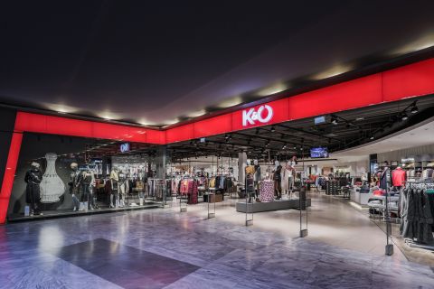 L'image montre l'extérieur d'un magasin de vêtements K&O dans un centre commercial. La façade du magasin est rouge et noire et présente des mannequins portant différentes tenues. À l'intérieur, on peut voir des portants à vêtements à côté d'un éclairage de plafond lumineux.