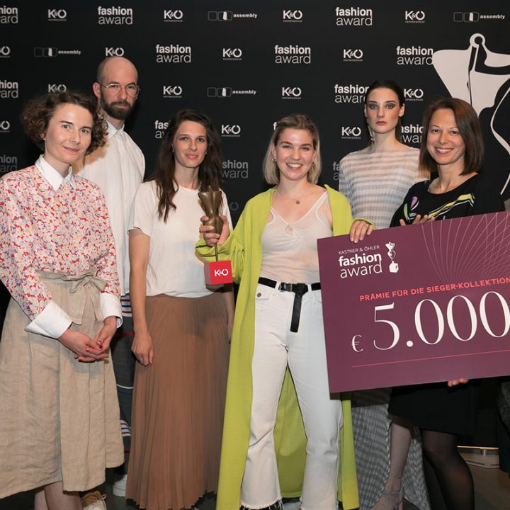 Un groupe de huit personnes pose lors d'un événement de mode. L'un d'eux tient un grand chèque de prix de 5 000 euros à la main. Ils se tiennent devant un fond noir avec des logos d'événements de mode. Certains portent des tenues élégantes et modernes.