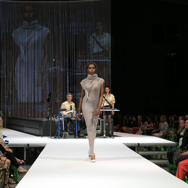 Un mannequin défile sur un podium lumineux, vêtu d'une tenue grise à col montant. À l'arrière-plan, un groupe avec un chanteur et un claviériste se produit. Le public est assis de part et d'autre de la scène et regarde le spectacle.