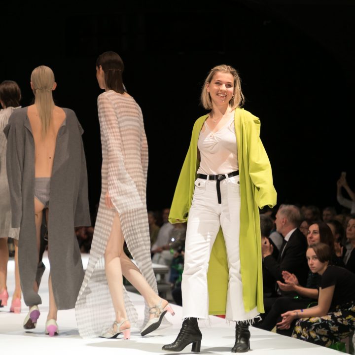 Un groupe de mannequins défile sur le podium dans des tenues claires et neutres. Une femme portant un manteau vert clair et un pantalon blanc les suit et sourit au public qui applaudit. Sous la scène, les gens regardent attentivement.