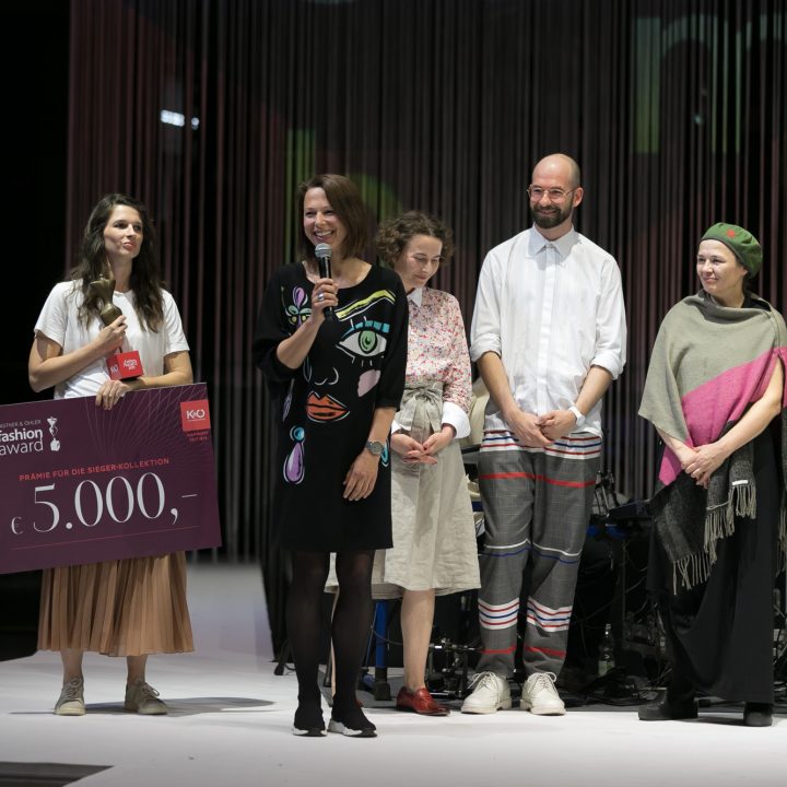 Un groupe de six personnes se tient sur une scène. Une femme tient un grand chèque de 5 000 euros à la main. Une autre femme parle dans un microphone. Les autres sourient. Tous portent des vêtements décontractés et colorés. Des spectateurs observent la scène de côté.