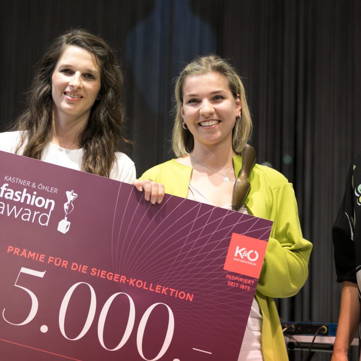 Trois femmes se tiennent côte à côte en souriant. La femme au milieu tient un grand chèque de 5 000 euros avec l'inscription « Kastner & Öhler Fashion Award ». Elles sont vêtues à la mode et semblent être lors d'un événement ou d'une cérémonie.