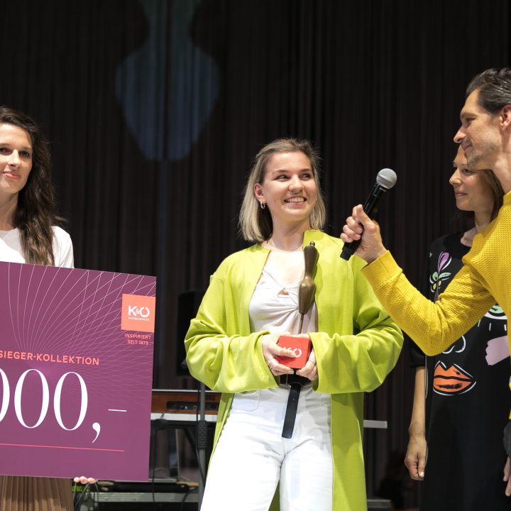Un groupe de trois femmes et un homme se tient sur scène. Une femme tient un grand chèque de 5 000 euros et un trophée à la main. L'homme avec le microphone parle. Ils semblent être à une remise de prix, il est écrit « Fashion Award » sur le chèque.