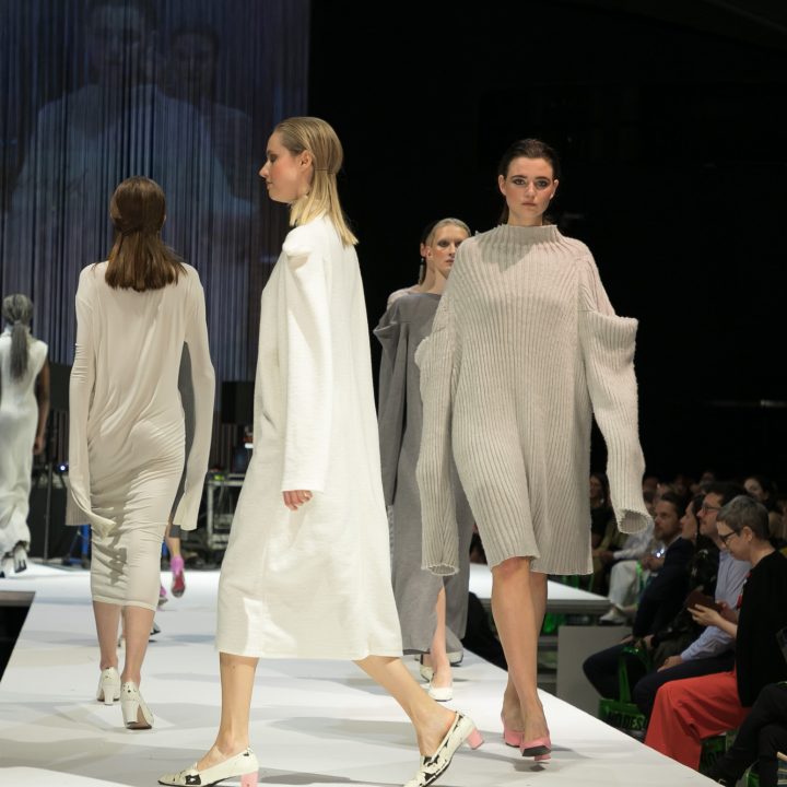 Des mannequins défilent sur le podium en robes en maille minimalistes surdimensionnées dans des tons neutres. Le public regarde des deux côtés dans une salle faiblement éclairée, avec le podium visible sur un grand écran en arrière-plan.
