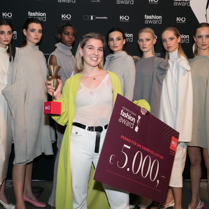 Une femme souriante tient un trophée et un grand chèque de 5 000 euros dans ses mains, devant un groupe de huit mannequins en robes de couleurs neutres. En arrière-plan, on aperçoit les logos d'une cérémonie de remise de prix de la mode.