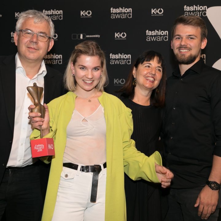 Quatre personnes sourient pour une photo lors d'une remise de prix de la mode. La femme au premier plan tient un trophée et porte un manteau vert clair. Les logos de l'événement sont visibles en arrière-plan.
