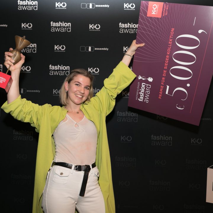 Une personne portant un manteau vert clair tient un trophée et un grand chèque de 5 000 euros dans ses mains lors d'une cérémonie de remise de prix de la mode. Les logos de l'événement et du sponsor sont visibles en arrière-plan. Elle sourit et semble excitée.