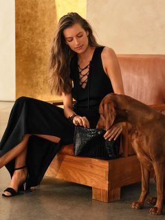 Une femme en robe noire est assise avec un chien marron sur un canapé marron et tient un sac à main noir sur ses genoux.