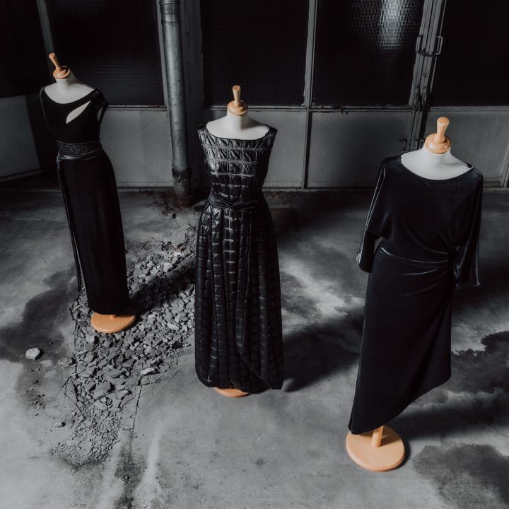Trois mannequins présentent d'élégantes robes de soirée noires. Chaque robe a son propre style et présente des textures et des coupes différentes. Ils se tiennent sur un sol en béton texturé dans un environnement industriel faiblement éclairé.