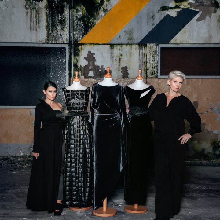 Deux femmes se tiennent à côté de trois mannequins portant d'élégantes robes noires. En arrière-plan, on aperçoit un mur industriel usé avec des motifs géométriques en jaune et gris. La scène dégage un mélange de mode moderne et d'esthétique urbaine.