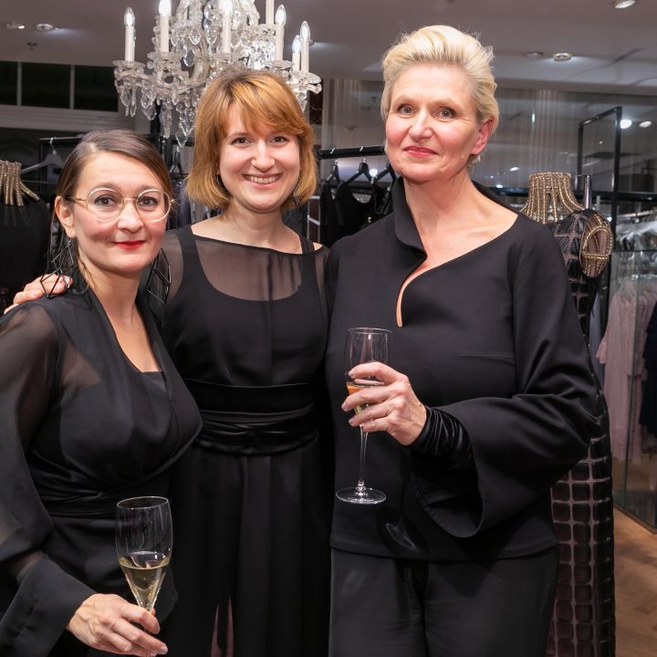 Trois femmes en tenues noires élégantes se tiennent ensemble, sourient et tiennent des coupes de champagne à la main. Elles se trouvent dans un magasin de vêtements. À l'arrière-plan, on aperçoit des portants à vêtements et un lustre.