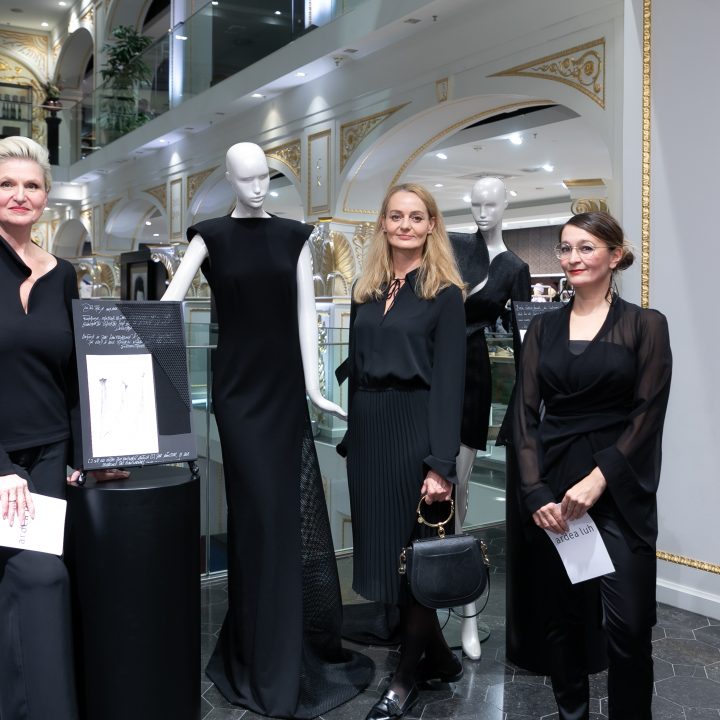 Trois femmes vêtues de noir se tiennent à côté de deux mannequins portant d'élégantes robes noires dans un intérieur stylé. Elles semblent être à un événement ou une exposition et tiennent des papiers dans leurs mains. À l'arrière-plan, on aperçoit des accents décoratifs dorés.