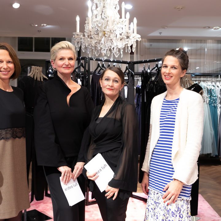 Quatre femmes se tiennent dans un magasin de vêtements chic et posent devant des robes élégantes exposées sur des portants. Un lustre est suspendu au-dessus d'elles, ajoutant une touche de raffinement à l'environnement. Deux femmes tiennent des brochures d'événements dans leurs mains.