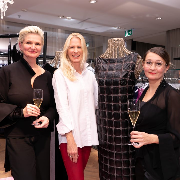 Trois femmes sourient, des coupes de champagne à la main, dans un magasin de vêtements. À côté d'elles se trouve un mannequin portant une robe à motifs. En arrière-plan, on aperçoit des portants à vêtements et un lustre au-dessus de leur tête.