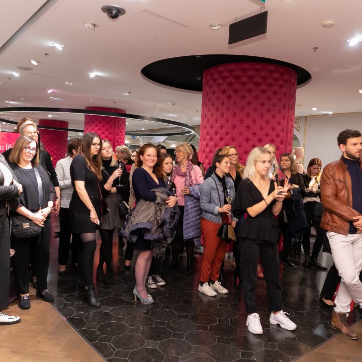 Un groupe de personnes se rassemble dans une pièce et regarde un événement ou une présentation. Ils sont vêtus de manière décontractée et se tiennent autour de tables avec des boissons. La pièce a des colonnes roses rondes et un décor moderne, créant une atmosphère animée.